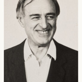 Homem branco, de meia idade, sorridente. Cabelos ralos e despenteados.
